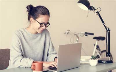 A woman using a laptop doing remote proctoring at a desk with a lamp.